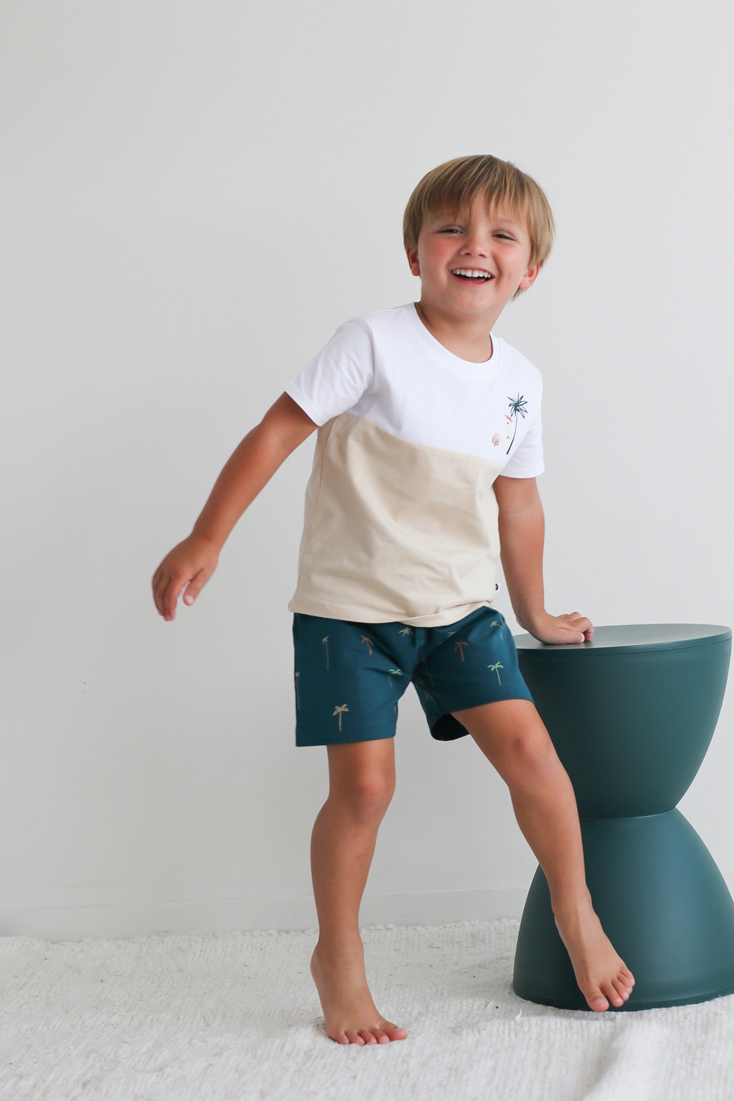 Boy leaning on a stool against a white background while wearing Jungle Stamp Block T-Shirt and Palm Leo Shorts, all designed by Burrow and Be