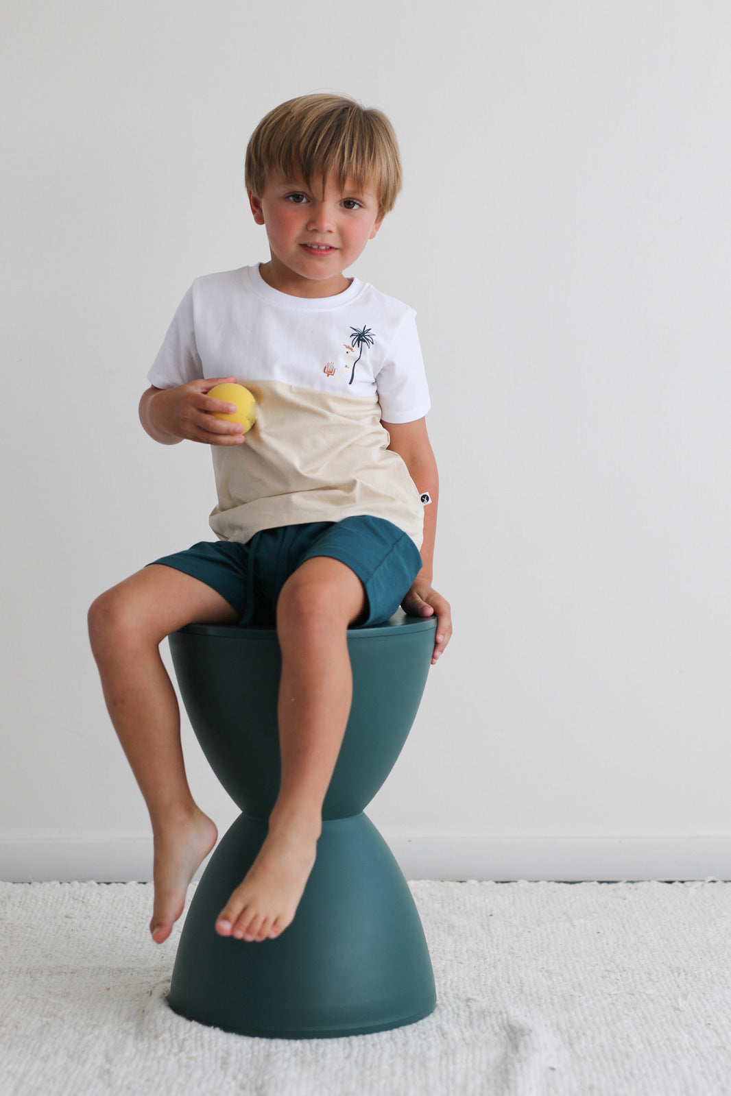 Boy holding a ball in hand and sitting on a stool against a white background while wearing Jungle Stamp Block T-Shirt and Leo Shorts in Deep Teal, all designed by Burrow and Be