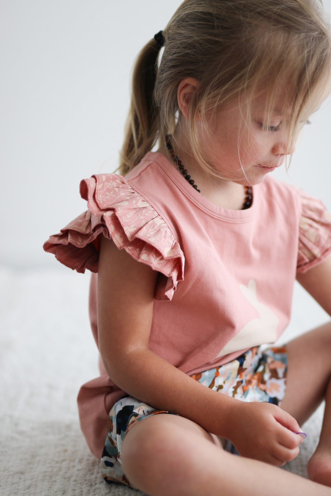 Girl sitting on white carpet floor wearing Doe Flutter T-shirt and Delight Lyla Shorts by Burrow and Be