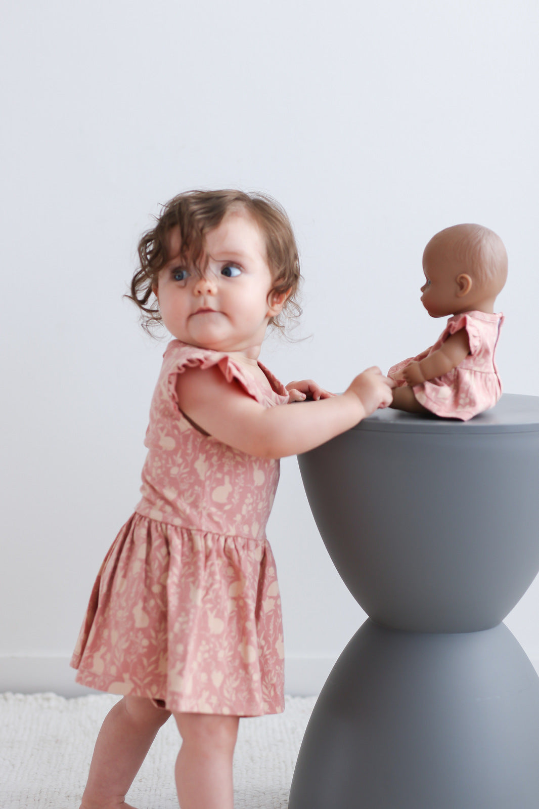 Baby standing in front of stool facing a doll, against a white background and wearing Mead-Doe Flutter Baby Dress, designed by Burrow and Be