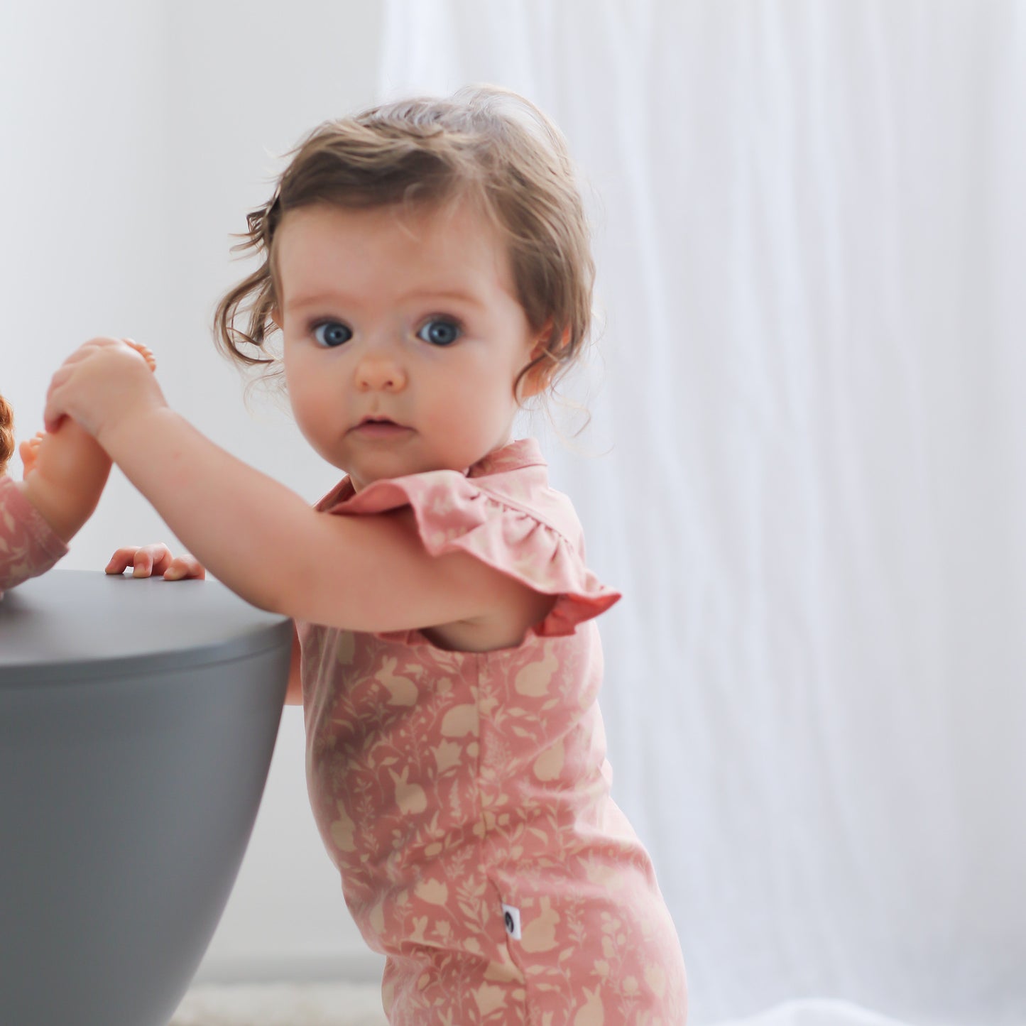Baby standing against a stool and wearing Mead-Doe Flutter Bodysuit, designed by Burrow and Be
