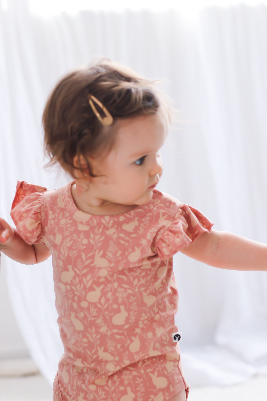 Baby girl posing in front of a white curtain and wearing Mead-Doe Flutter Bodysuit, designed by Burrow and Be