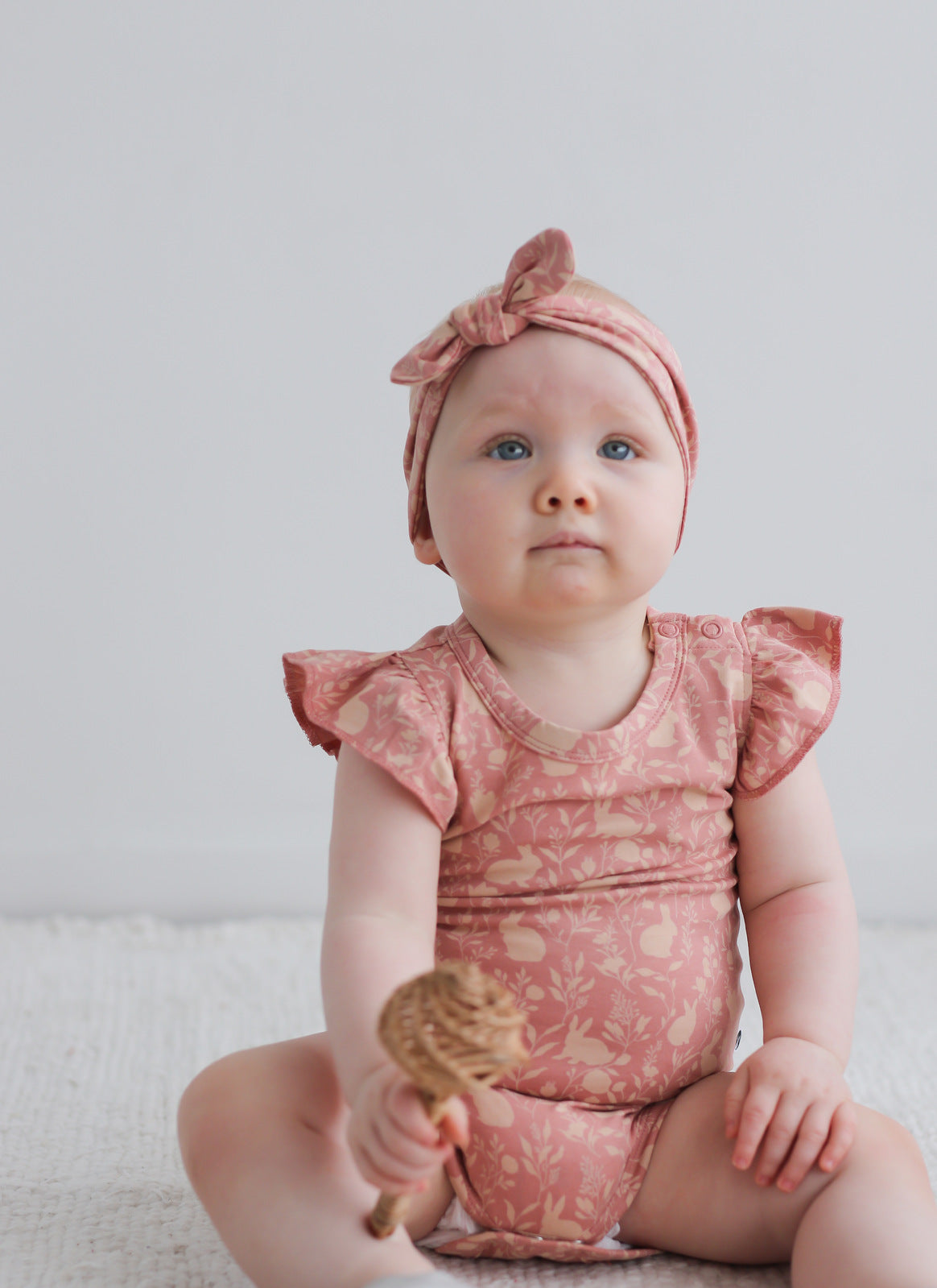 Baby girl sitting on white carpet and holding a wooden toy, while wearing Mead-Doe Headband and Flutter Bodysuit, all designed by Burrow and Be
