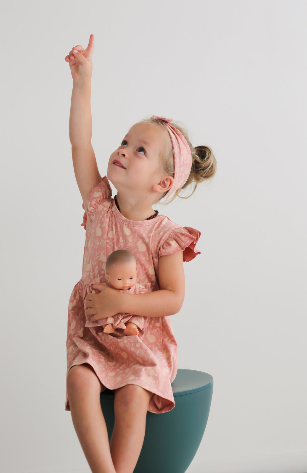 Girl sitting on a stool in front of a white wall, carrying a doll while wearing Mead-Doe Headband and Flutter Dress, all designed by Burrow and Be