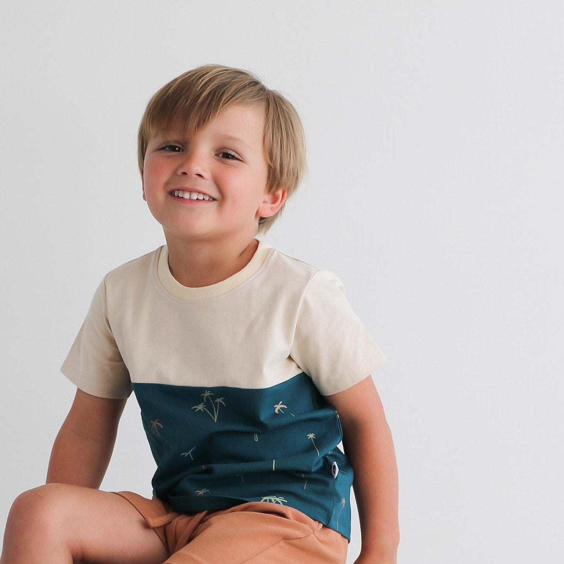 Close up of boy sitting on stool against a white background, wearing Palm Block T-shirt and Leo Shorts in Tan, both designed by Burrow and Be