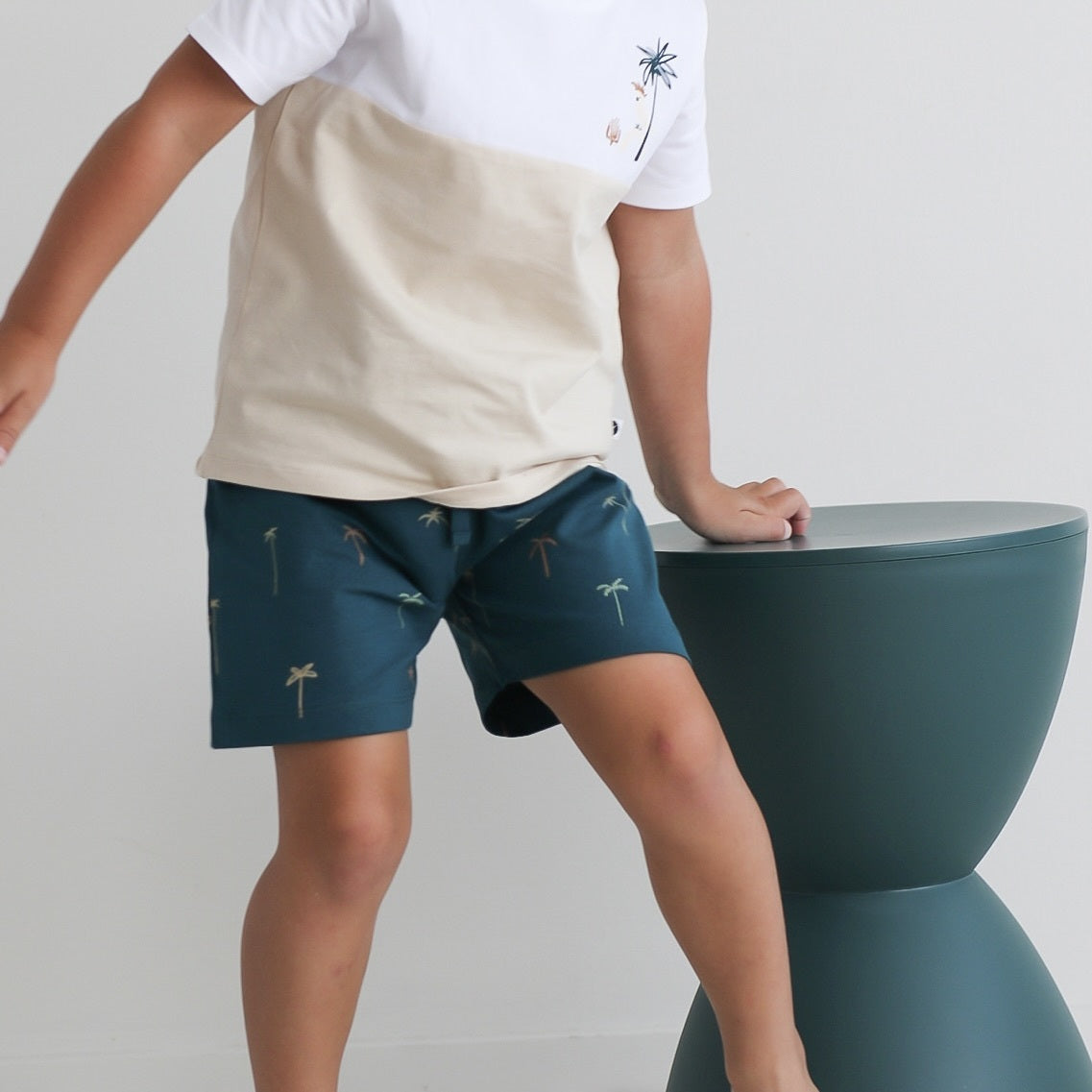Close up shot of boy leaning on a stool against a white background while wearing Jungle Stamp Block T-Shirt and Palm Leo Shorts, all designed by Burrow and Be
