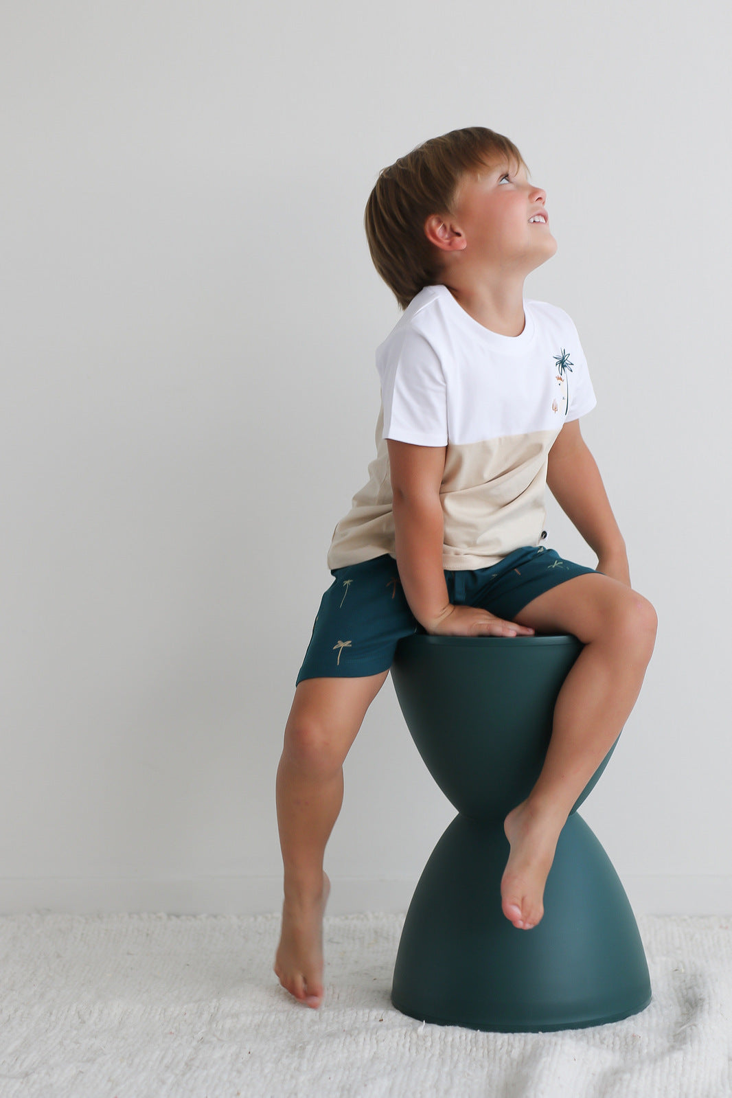 Boy sitting on a stool against a white background while wearing Jungle Stamp Block T-Shirt and Palm Leo Shorts, all designed by Burrow and Be