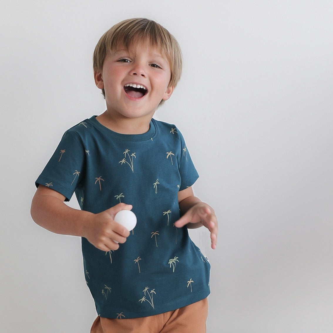 Close up of boy holding a ball in front of a stool against a white background, wearing Palm Classic T-shirt and Leo Shorts in Tan, both designed by Burrow and Be