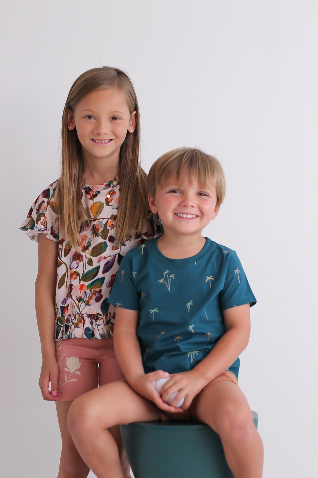 Girl wearing Garden Party Ella Top and Doe Bike Shorts and boy wearing Palm Classic T-shirt and Leo Shorts in Tan, all designed by Burrow and Be. Both posing against a white background,