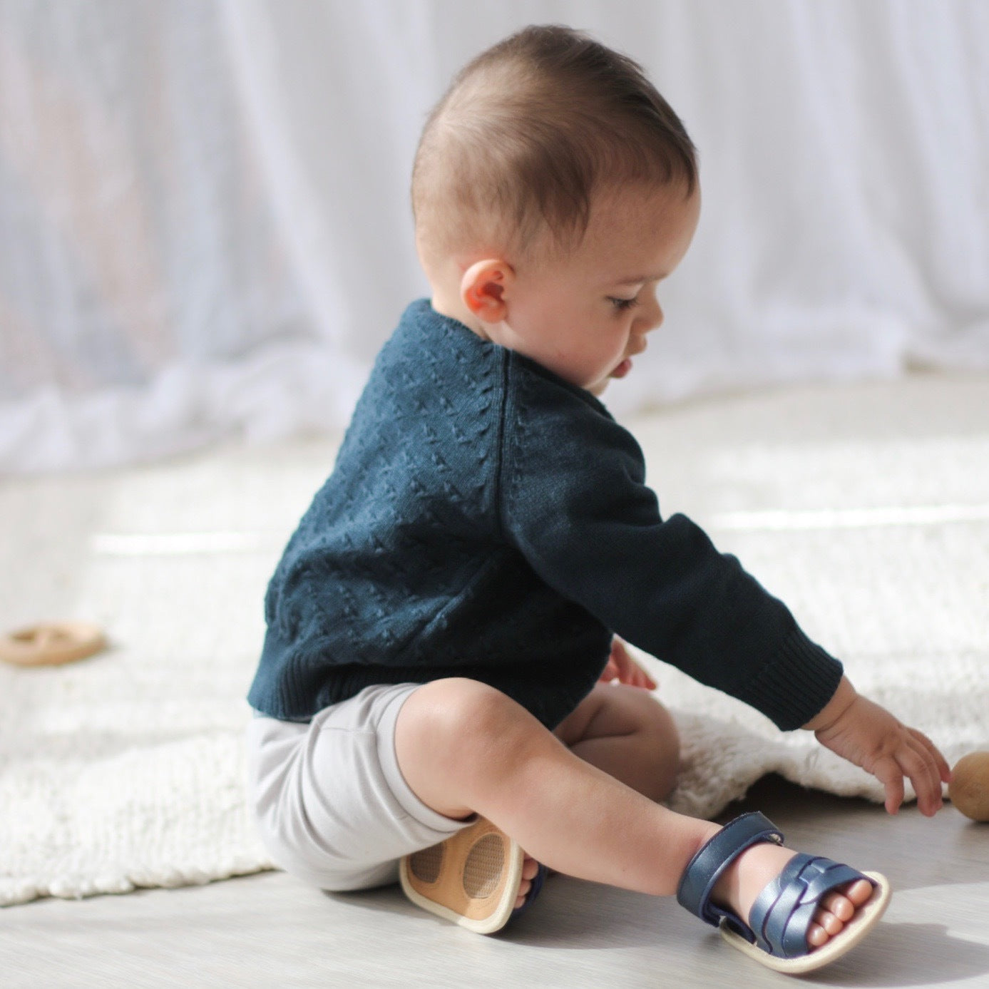 Baby sitting on floor against white curtain while wearing Mist baby pants andBillie Knit Cardigan by Burrow and Be and Sandals from Wander series