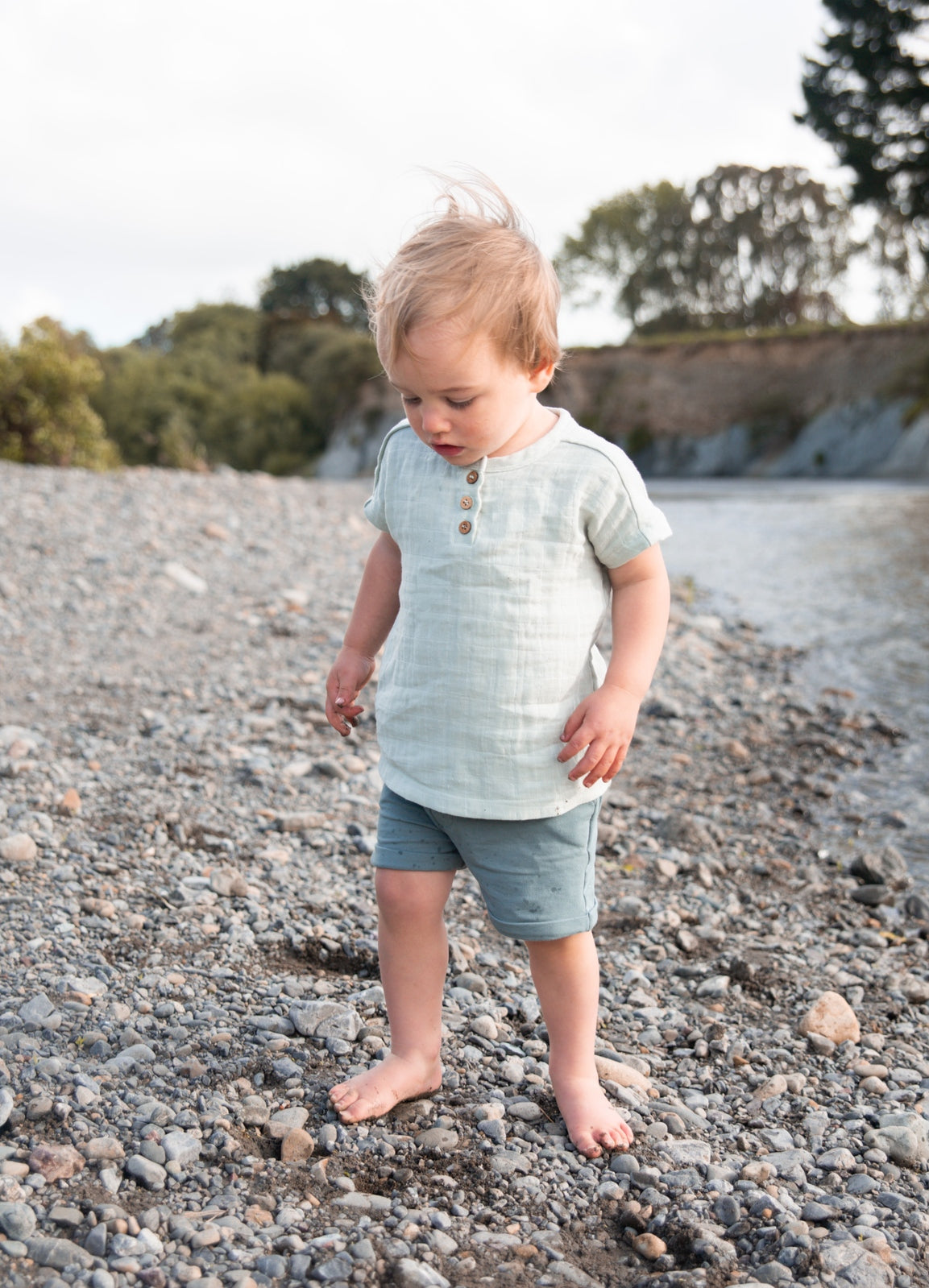 Boy standing on a rocky river bank, posing in wearing Jasper Muslin Top in Sage and Terry Knit Baby Shorts in Storm, both designed by Burrow and Be.