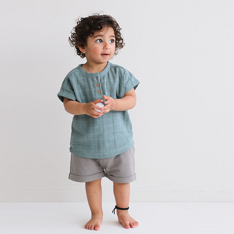 Boy standing in front of a white background, posing in wearing Jasper Muslin Top in Storm and Terry Knit Baby Shorts in Steel, both designed by Burrow and Be.