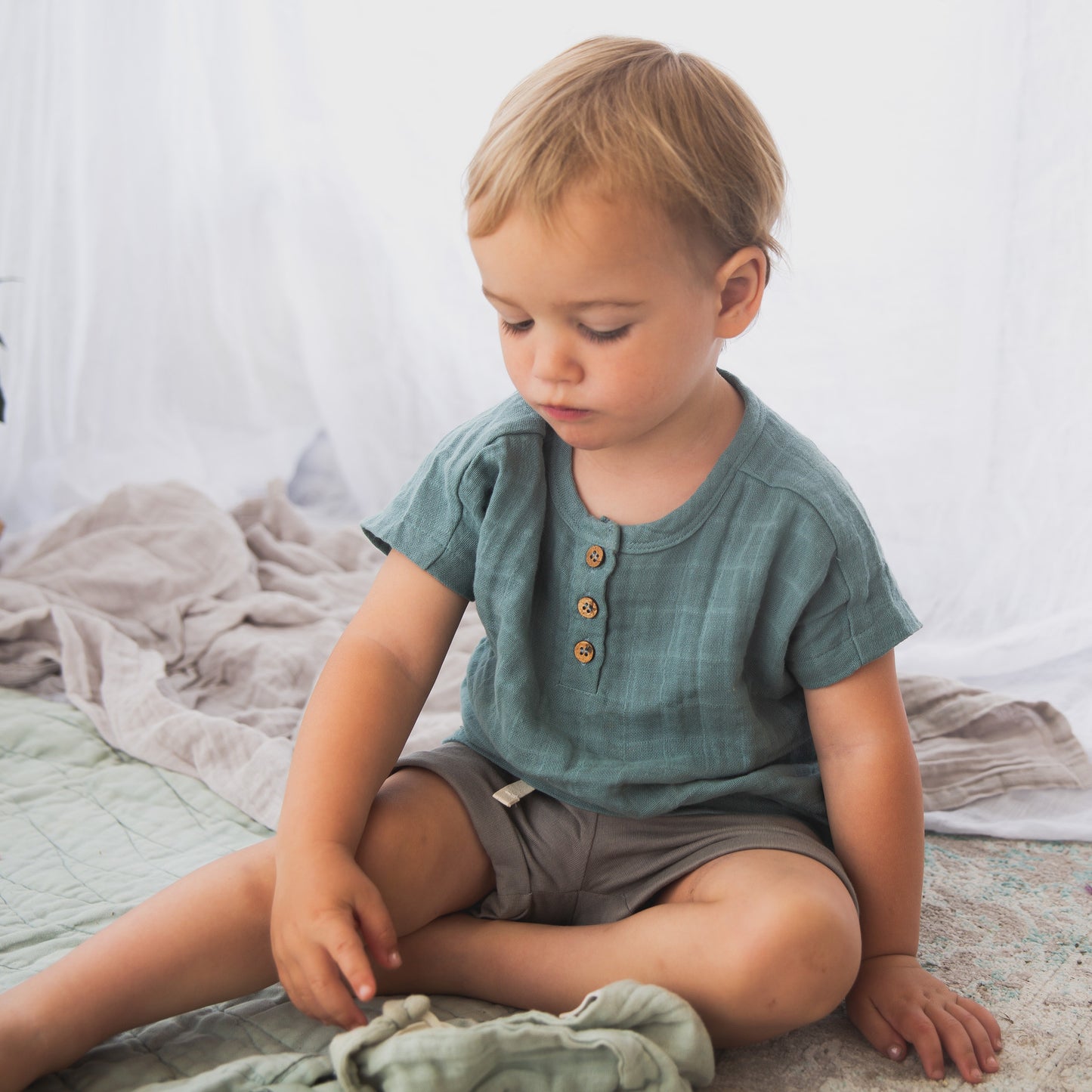 Boy posing against a white curtain while wearing a Jasper Muslin Top in Storm and Terry Knit Baby Shorts in Steel, both designed by Burrow and Be.