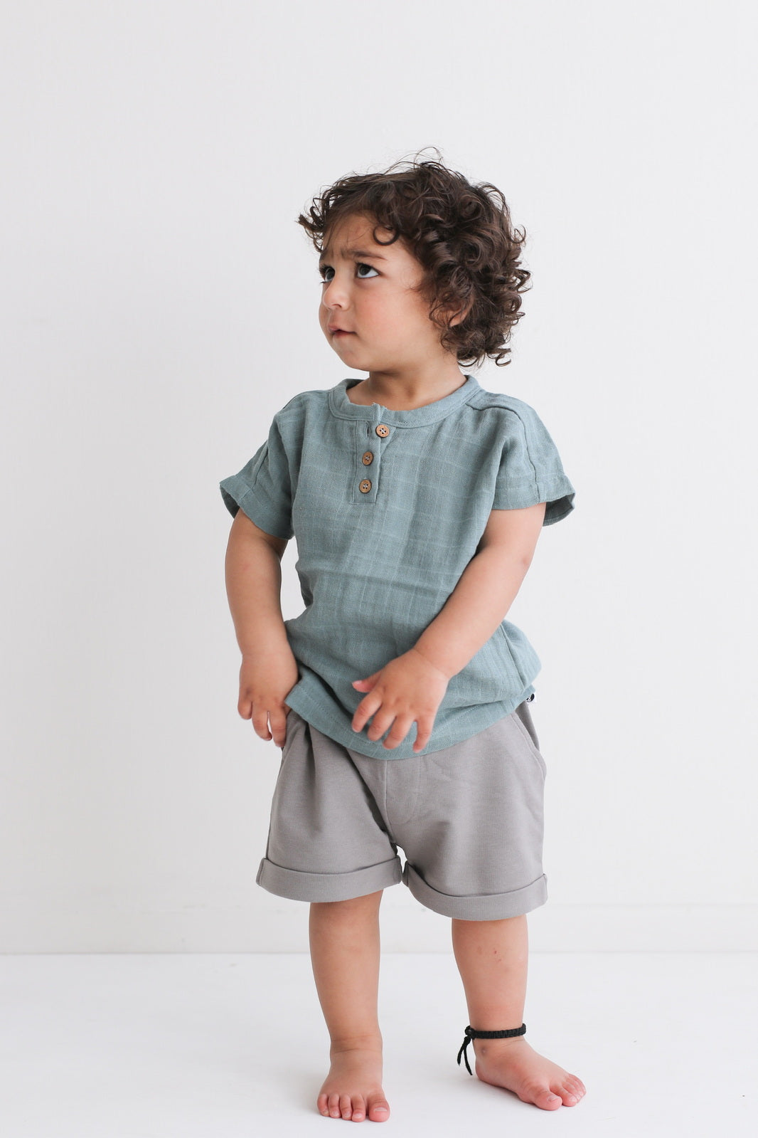 Boy standing in front of a white background looking into the distance, posing in wearing Jasper Muslin Top in Storm and Terry Knit Baby Shorts in Steel, both designed by Burrow and Be.