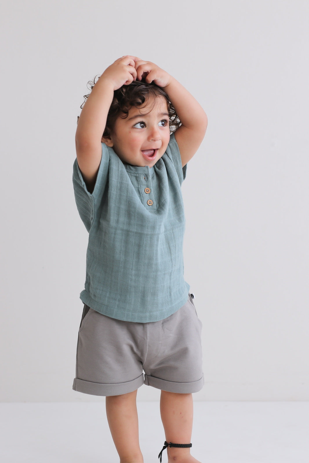 Boy posing with Jasper Muslin Top in Storm and Terry Knit Baby Shorts in Steel, both designed by Burrow and Be, against a white background
