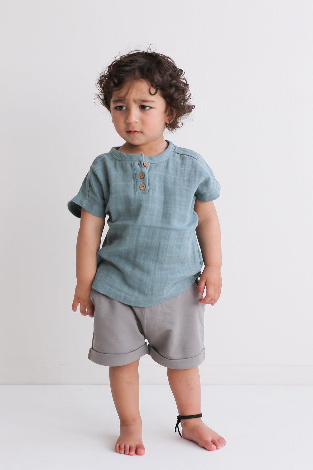Boy standing in front of a white background, posing in wearing Jasper Muslin Top in Storm and Terry Knit Baby Shorts in Steel, both designed by Burrow and Be.