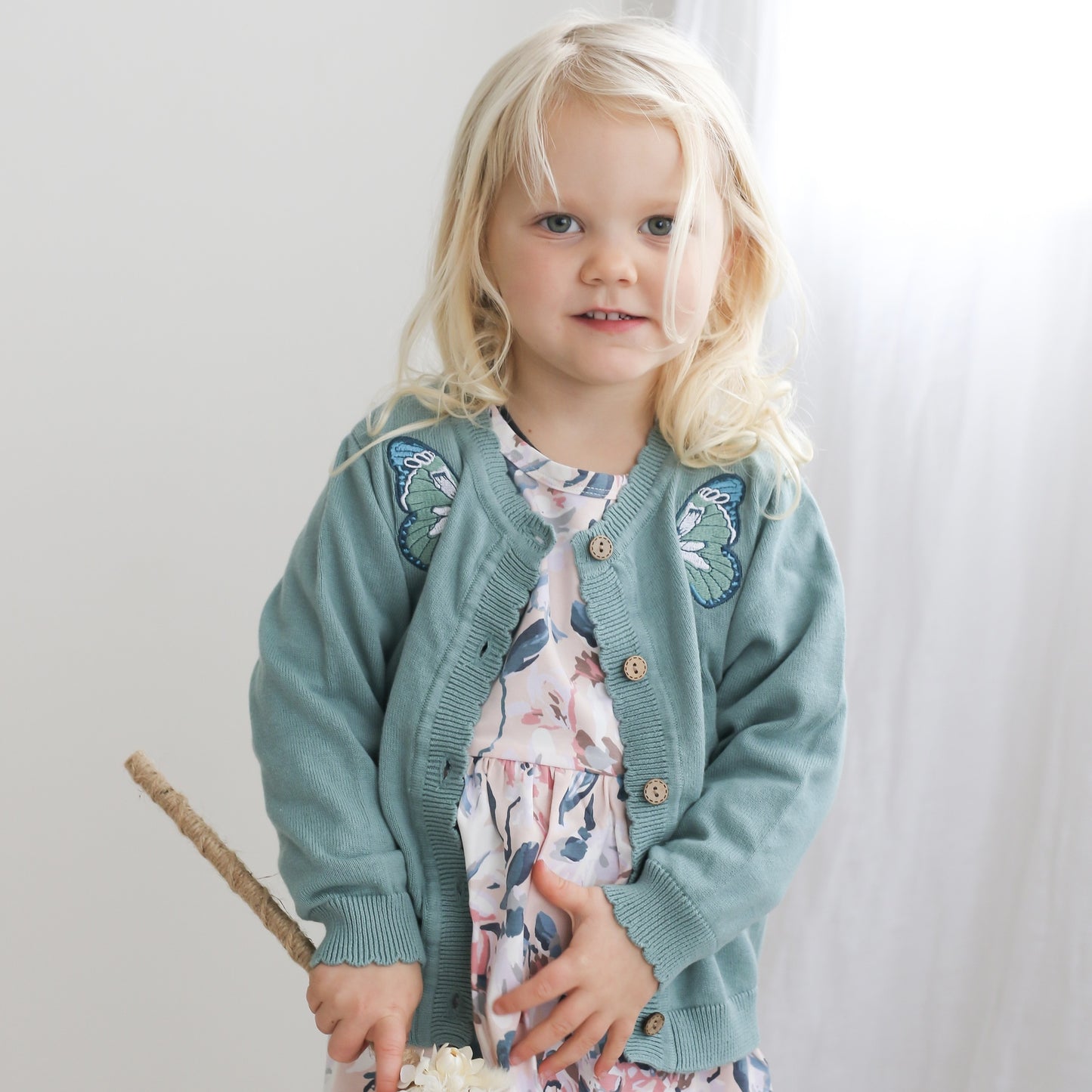Girl standing in front of a white background and white curtain while wearing Lola Knit Cardigan - Butterfly, designed by Burrow and Be