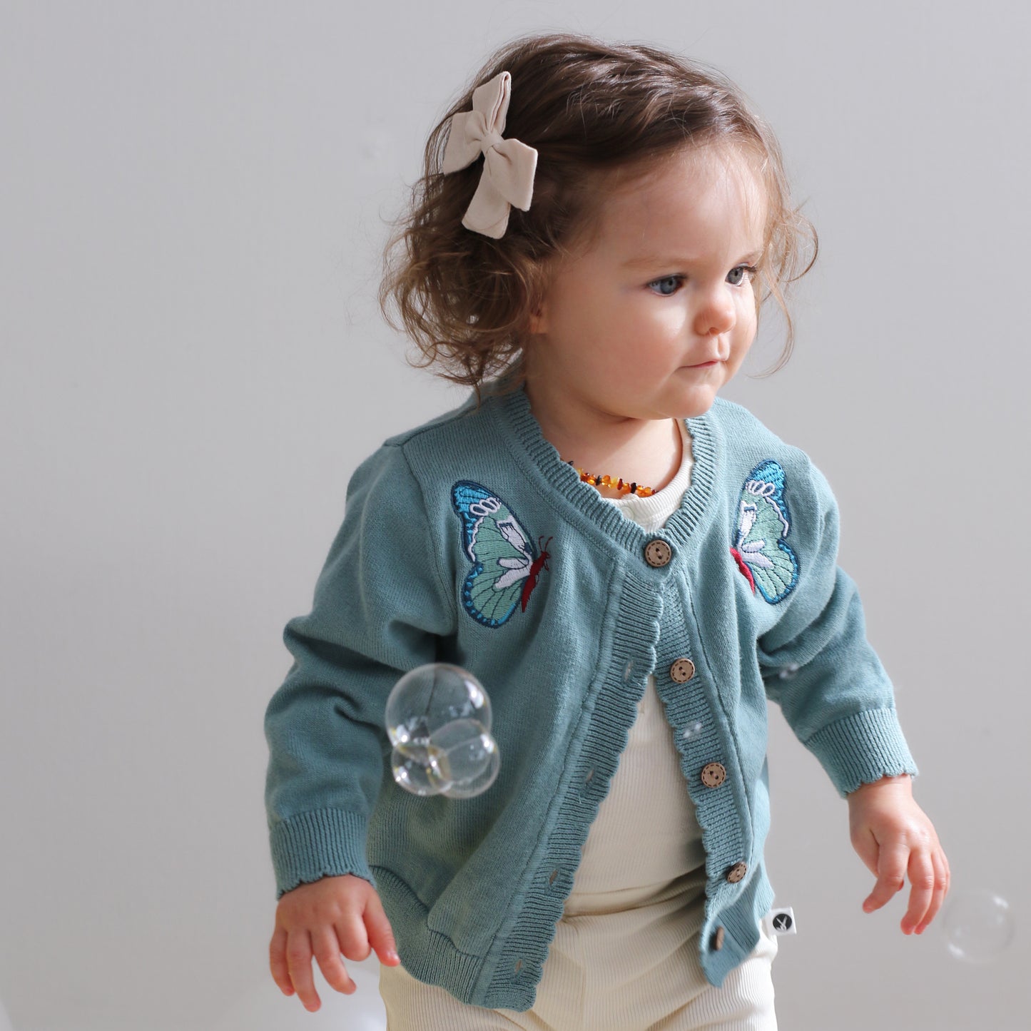 Baby girl playing with bubbles against a white background, wearing a Bow Tie hair clip, Bodysuit and Leggings all in Dusty Rose and Lola Knit Cardigan - Butterfly, all designed by Burrow and Be