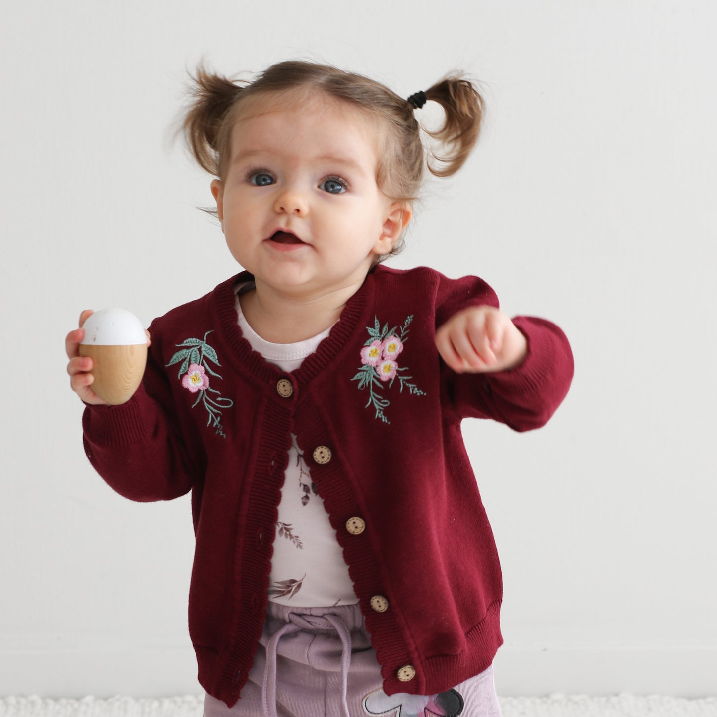 Girl holding a wooden toy, standing in front of a white background and wearing Lola Knit Cardigan - Bloom, designed by Burrow and Be
