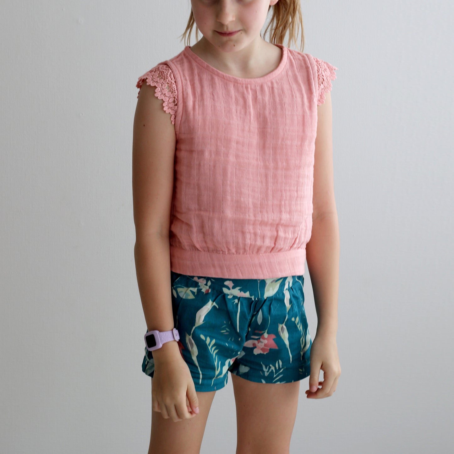 Girl standing in front of a white wall while wearing Dusty Rose Laced-Sleeve Woven Top and Green Leavings Lyla Shorts, all designed by Burrow and Be