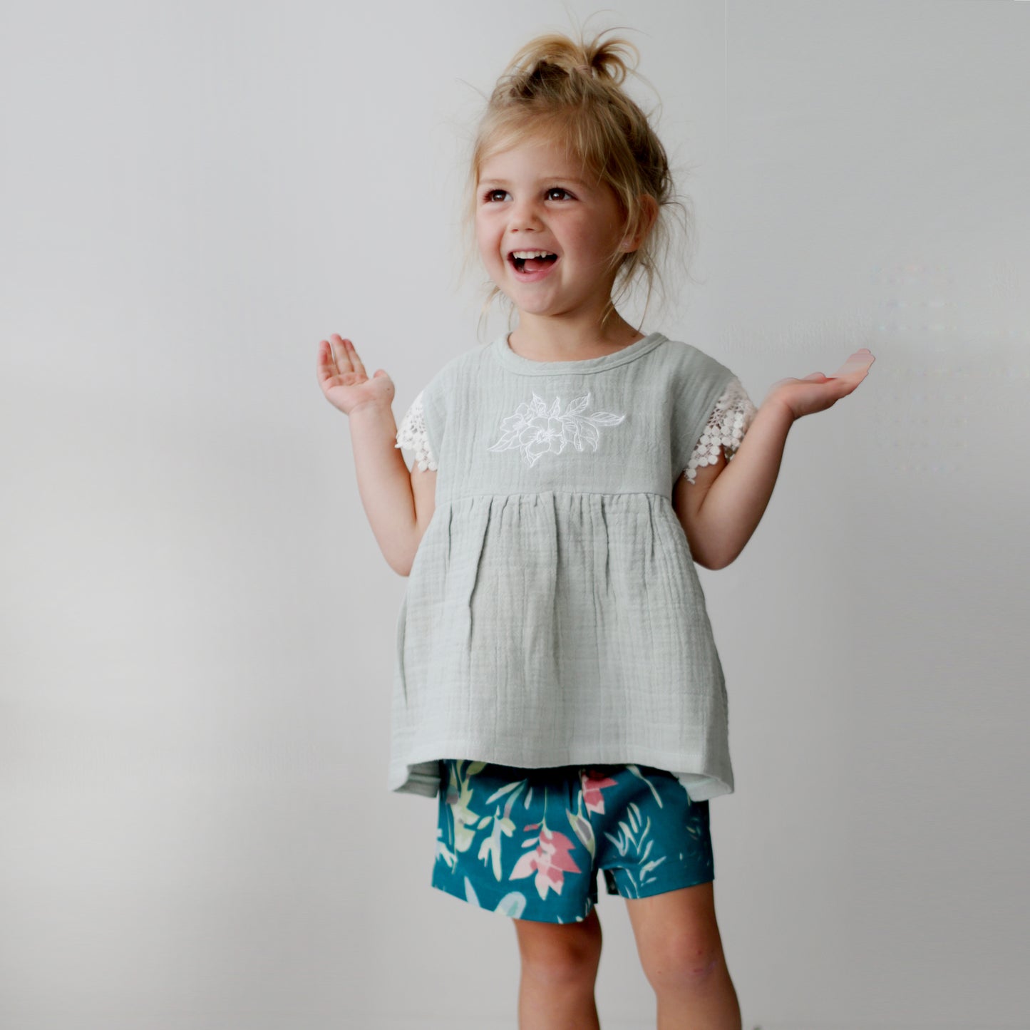 Girl standing in front of a white wall while wearing a Sage Woven Top and Green Leavings Lyla Shorts, all designed by Burrow and Be
