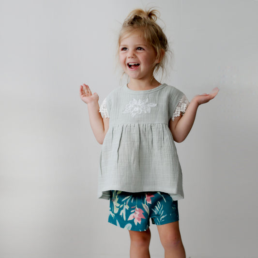Girl standing in front of a white wall while wearing a Sage Woven Top and Green Leavings Lyla Shorts, all designed by Burrow and Be
