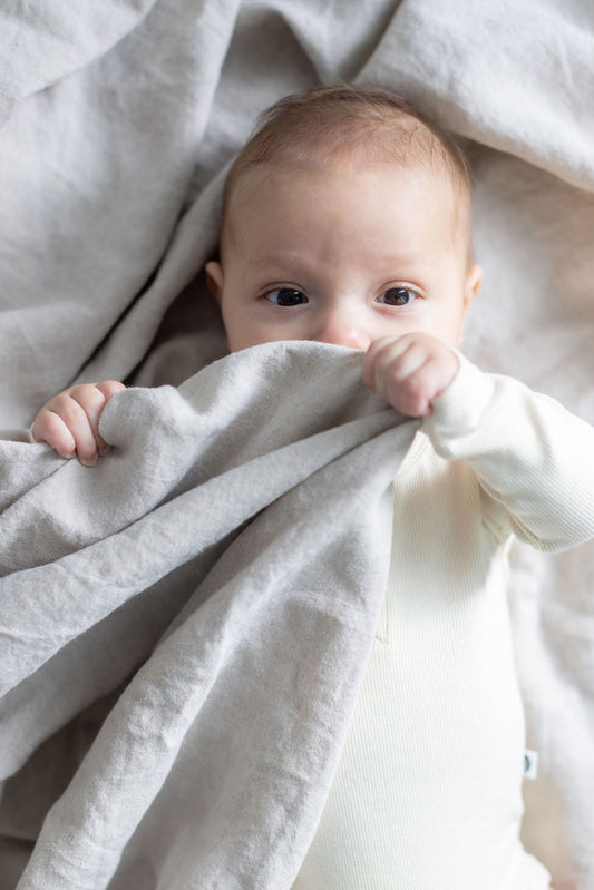 Baby in cream organic cotton bodysuit holding soft linen blanket, illustrating safe fabric-to-skin contact, Burrow and Be Singapore