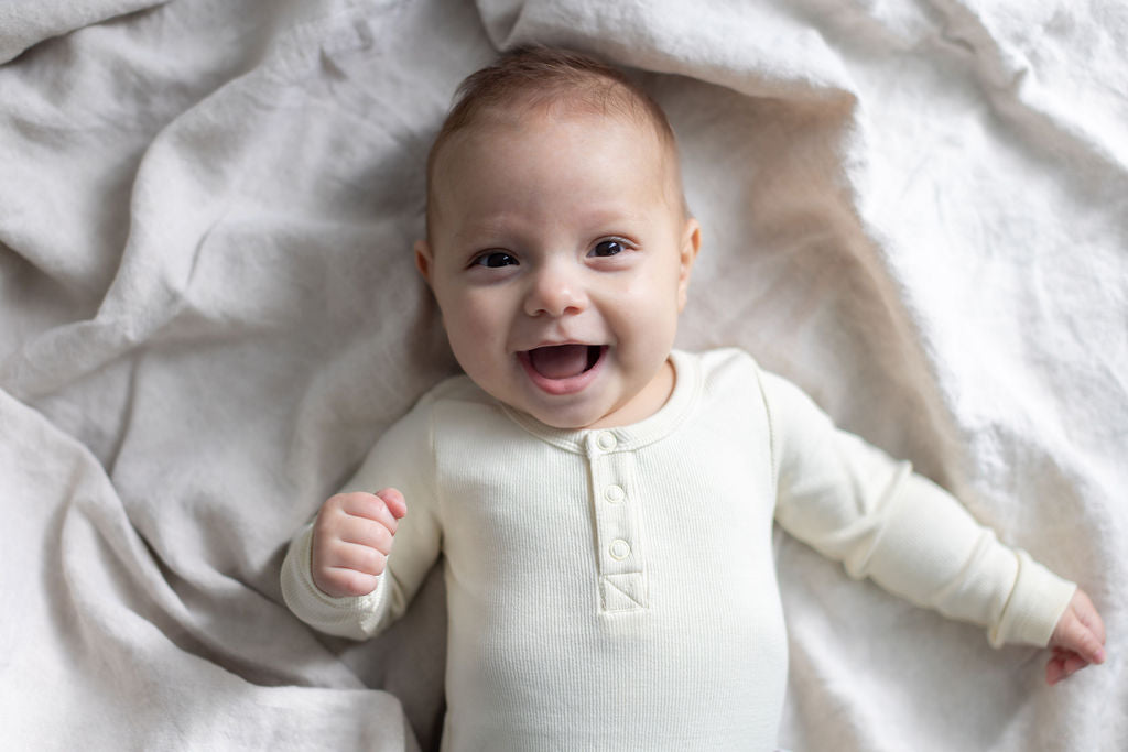 Wide-eyed laughing baby in cream organic cotton bodysuit on natural linen, illustrating curious confident development in the first year, Burrow and Be Singapore