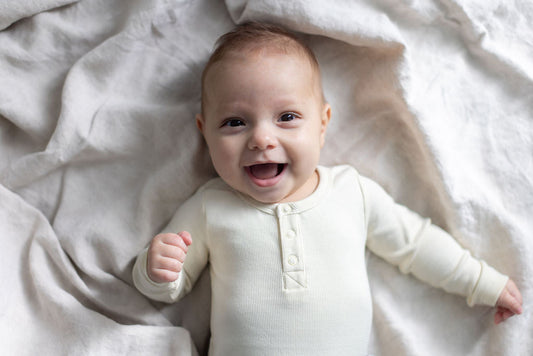Wide-eyed laughing baby in cream organic cotton bodysuit on natural linen, illustrating curious confident development in the first year, Burrow and Be Singapore
