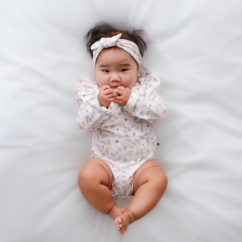 Infant wearing a white long-sleeve floral organic cotton romper and matching headband lying on a soft white bed