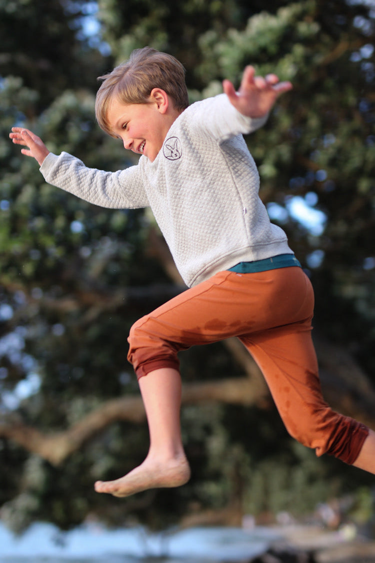 Active toddler jumping in the air wearing a grey quilted organic cotton sweater with a rabbit logo and rust-colored trousers