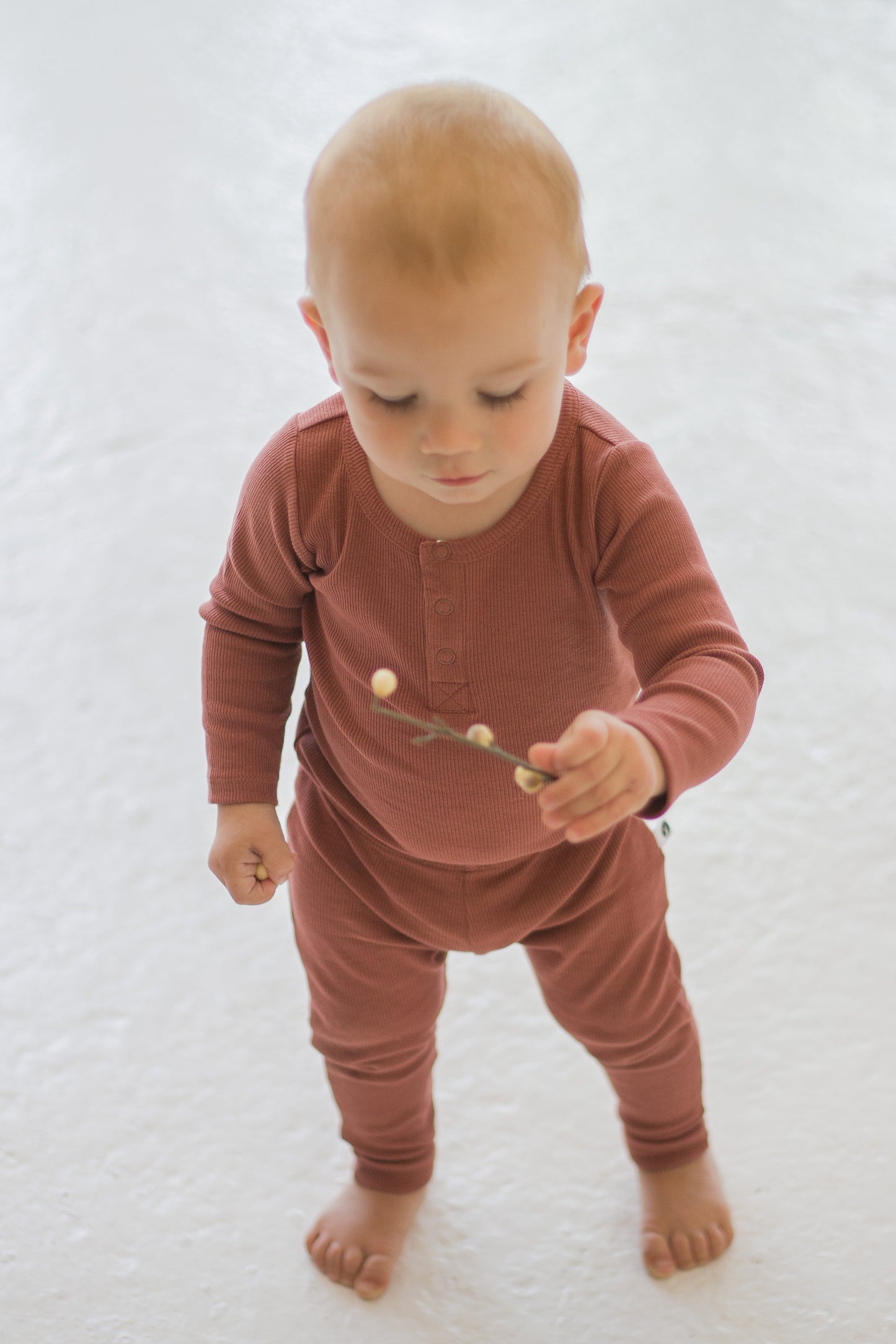 Boy standing on white carpet against a white background while wearing a Long Sleeve Henley Rib Bodysuit and Leggings in Clay, designed by Burrow and Be