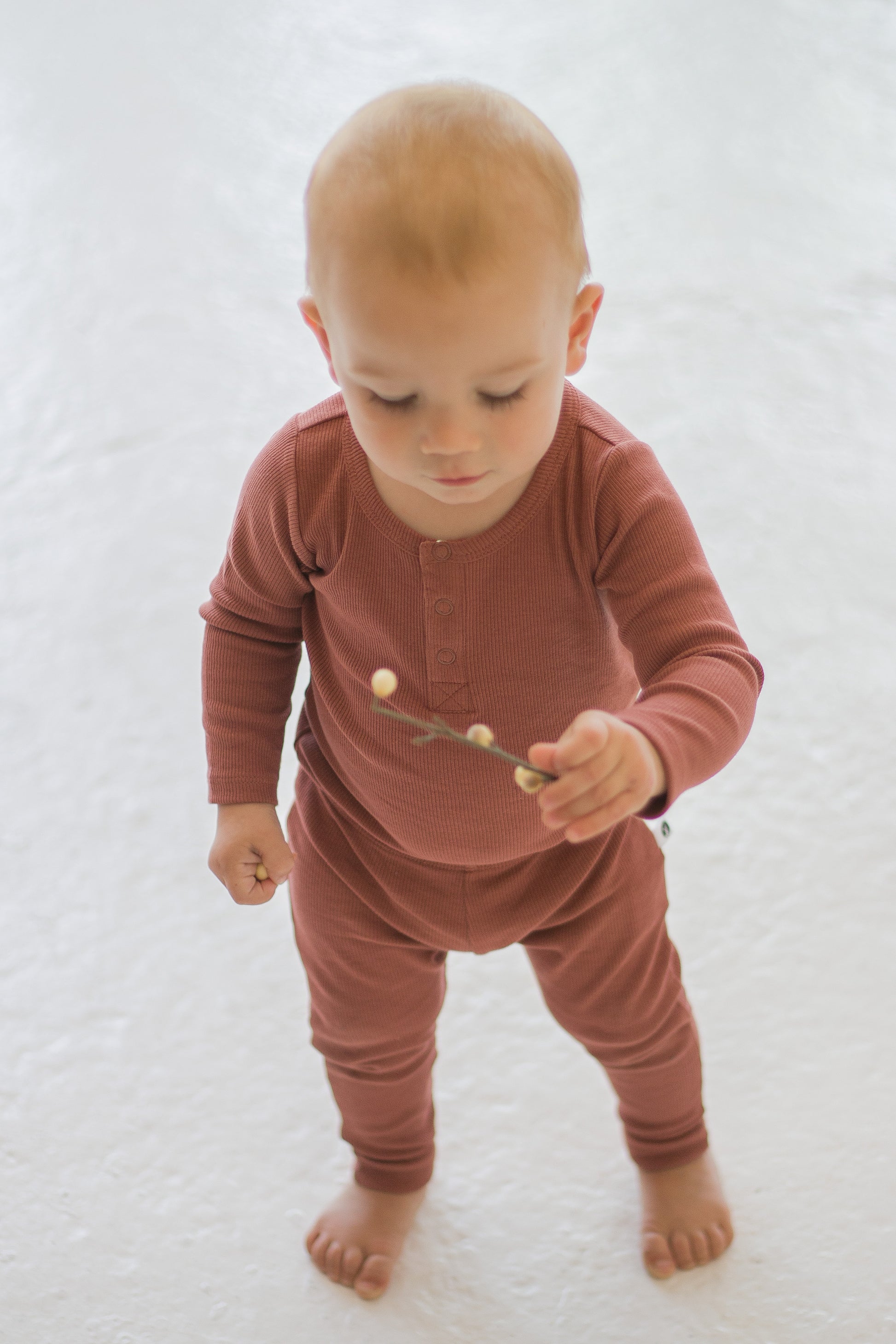 Boy standing on white carpet against a white background while wearing a Long Sleeve Henley Rib Bodysuit and Leggings in Clay, designed by Burrow and Be
