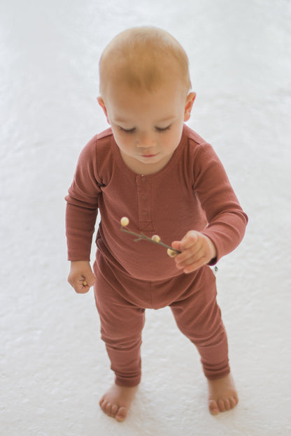 Boy standing on white carpet against a white background while wearing a Long Sleeve Henley Rib Bodysuit and Leggings in Clay, designed by Burrow and Be
