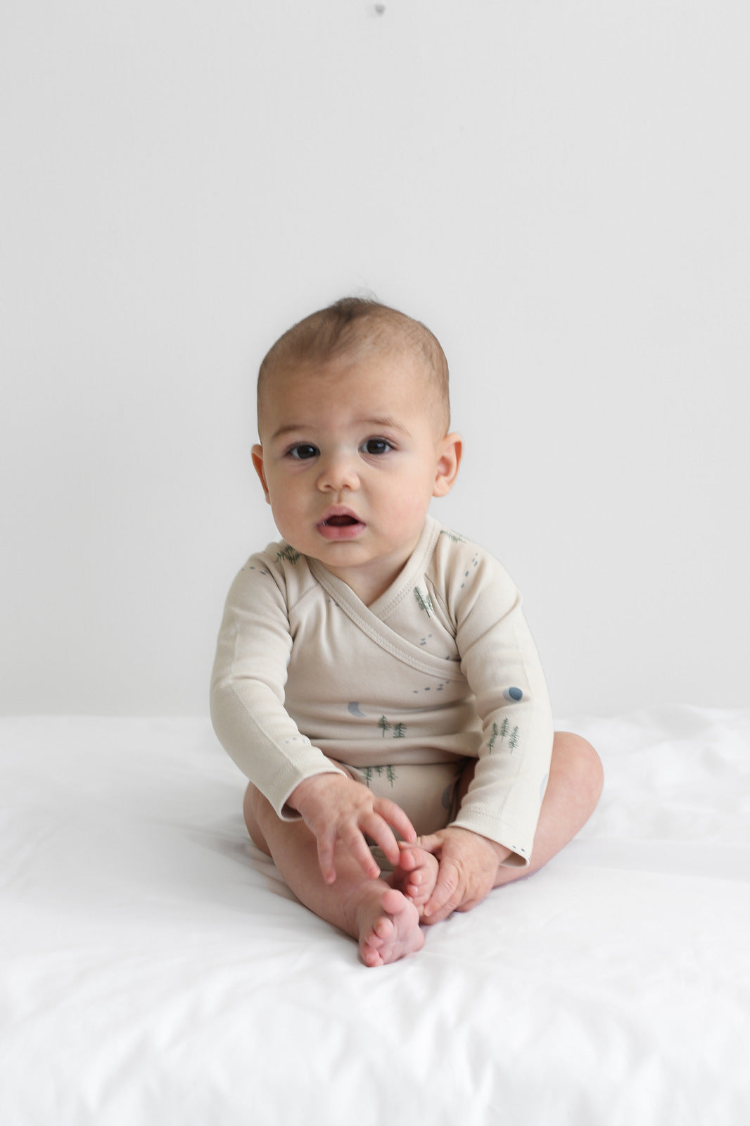 Baby sitting on white sheets in front of a white wall, while wearing Midnight Forest Kimono Bodysuit, designed by Burrow and Be