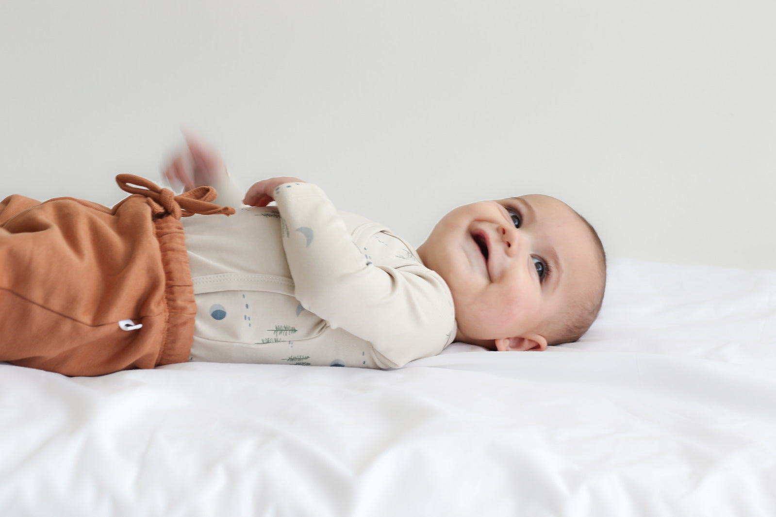 Baby laying on white sheets in front of a white wall, while wearing Leo Shorts in Tan and Midnight Forest Kimono Bodysuit, all designed by Burrow and Be