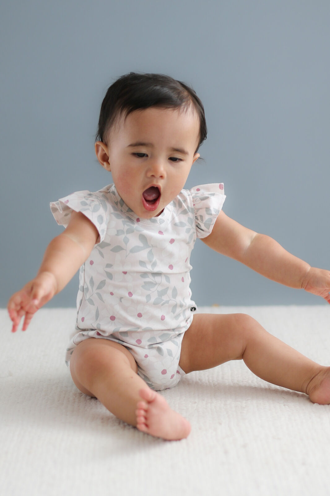 Baby sitting on a white surface wearing a patterned onesie against a gray background