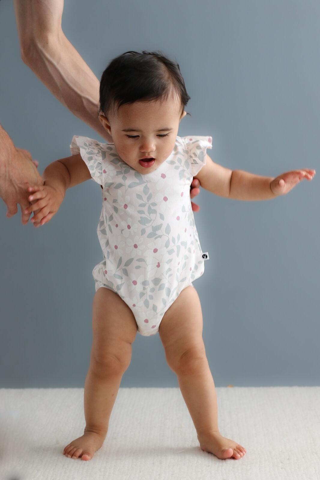 Baby in a white onesie with ruffled sleeves standing on a light surface against a gray background