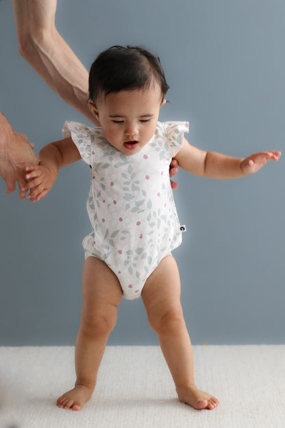 Baby in a white onesie with ruffled sleeves standing on a light surface against a gray background