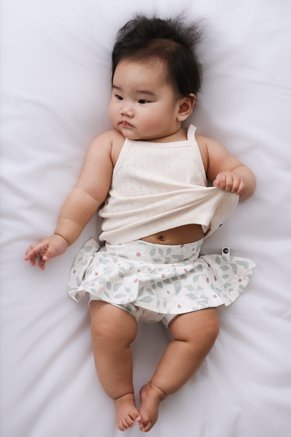 Baby lying on a white surface wearing a light-colored top and patterned skirt.
