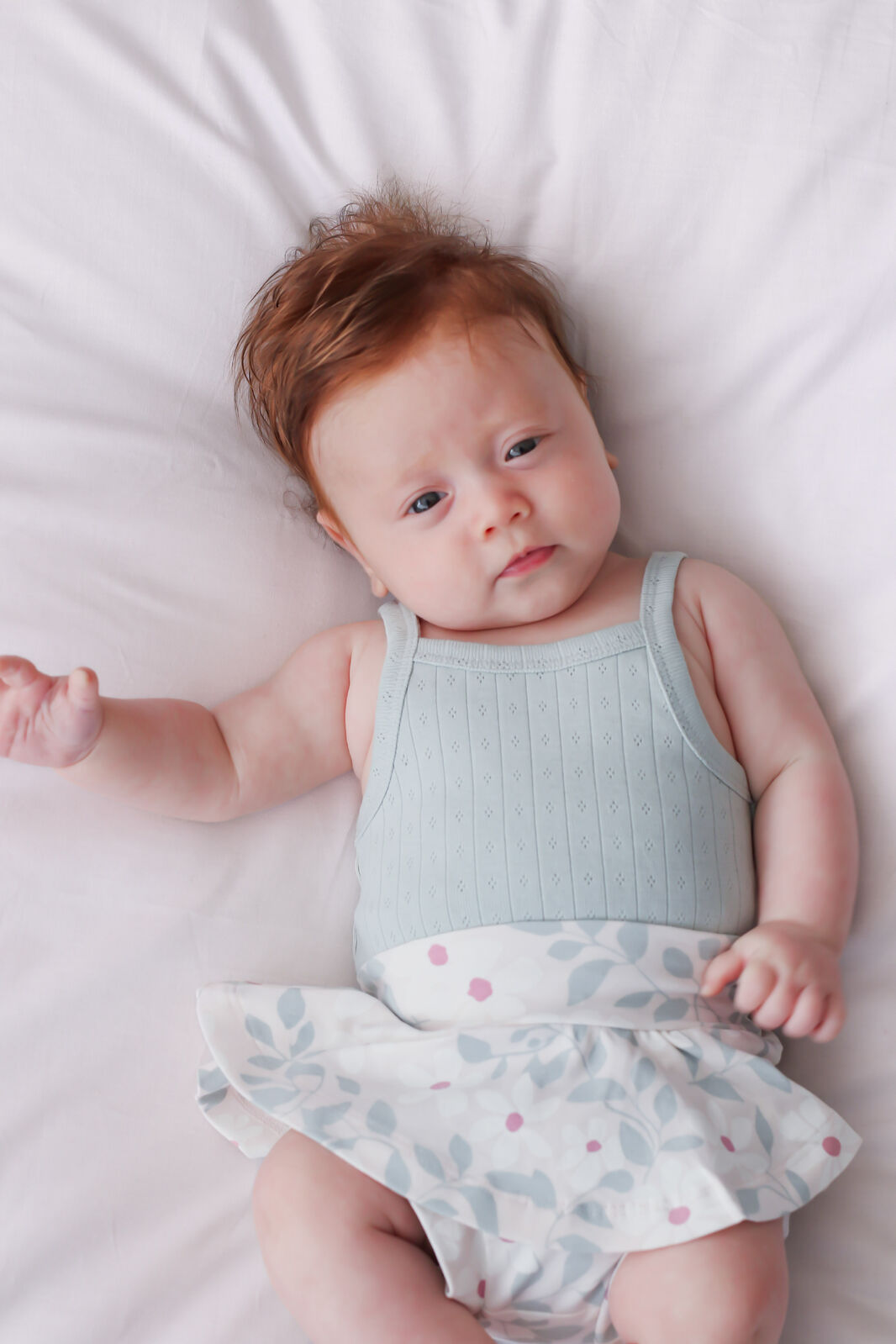 Baby lying on a white surface wearing a light blue sleeveless top and patterned skirt.