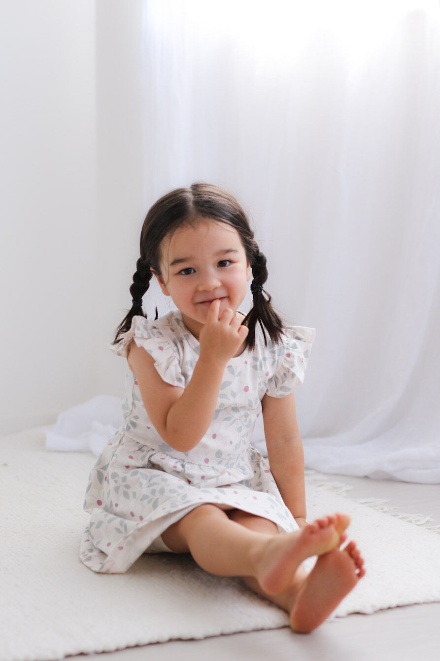 Young girl in a floral dress sitting on a white surface with a white curtain background