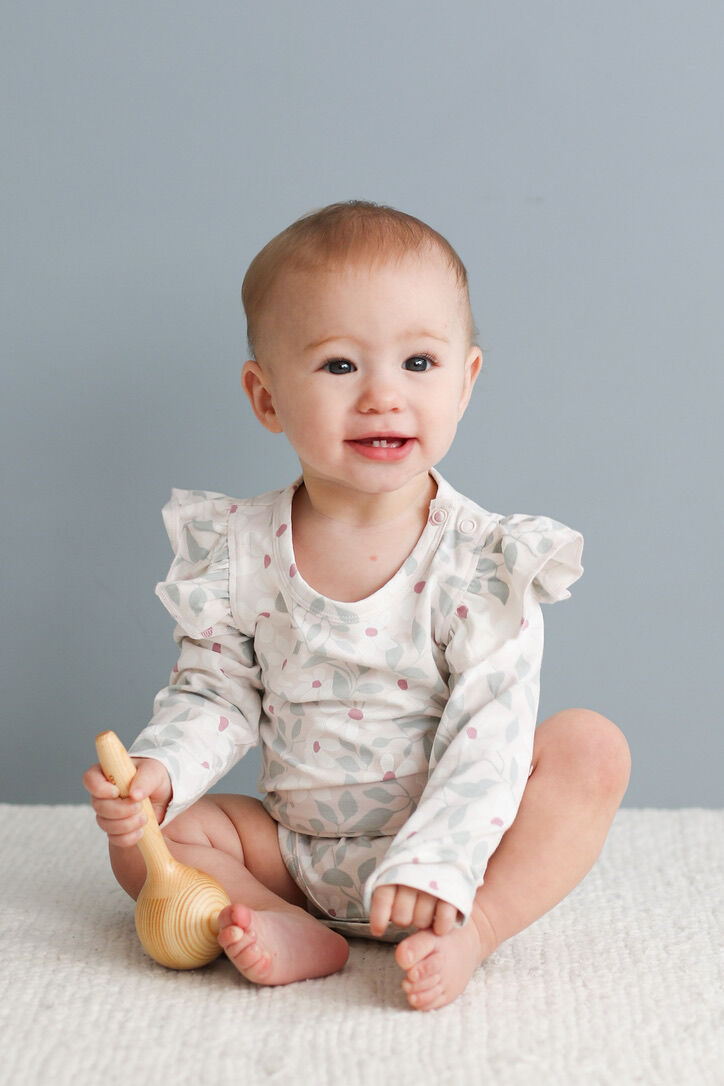 Baby sitting on a white blanket with a wooden toy, wearing a white romper with ruffles against a gray background.