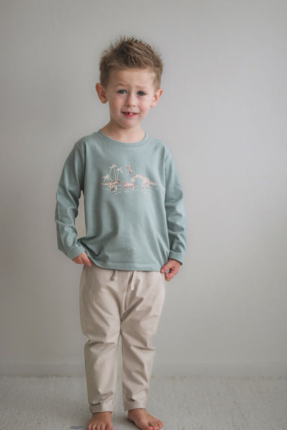 Young boy wearing a light green long-sleeve shirt and beige pants against a plain background