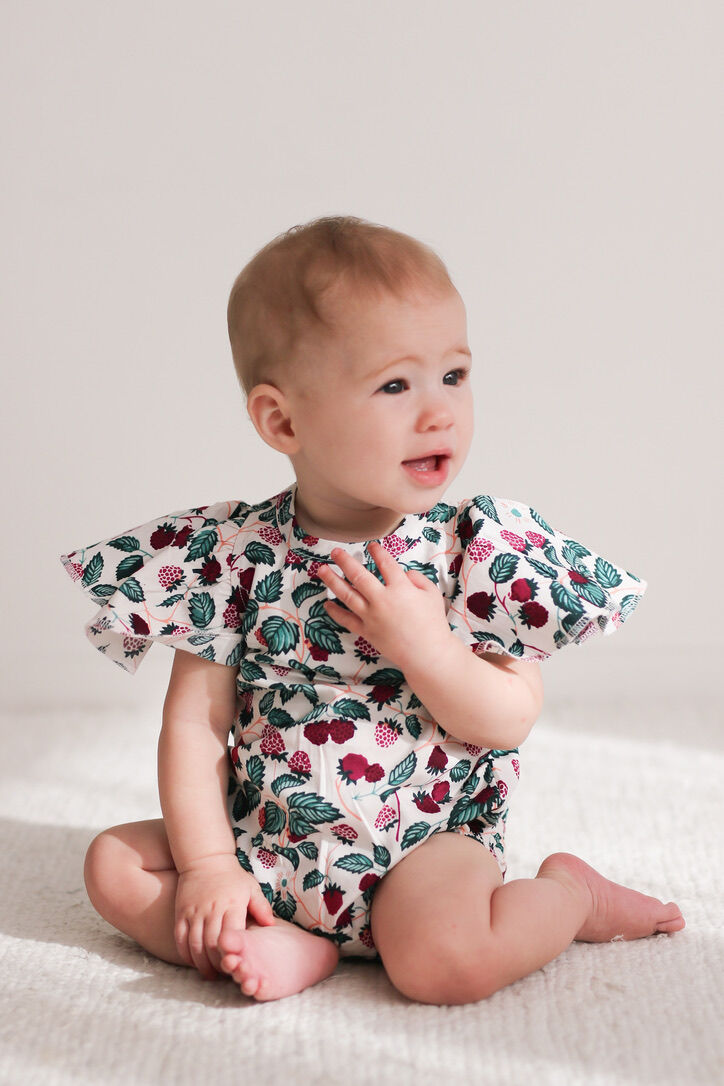 Baby wearing a floral romper sitting on a light-colored blanket.