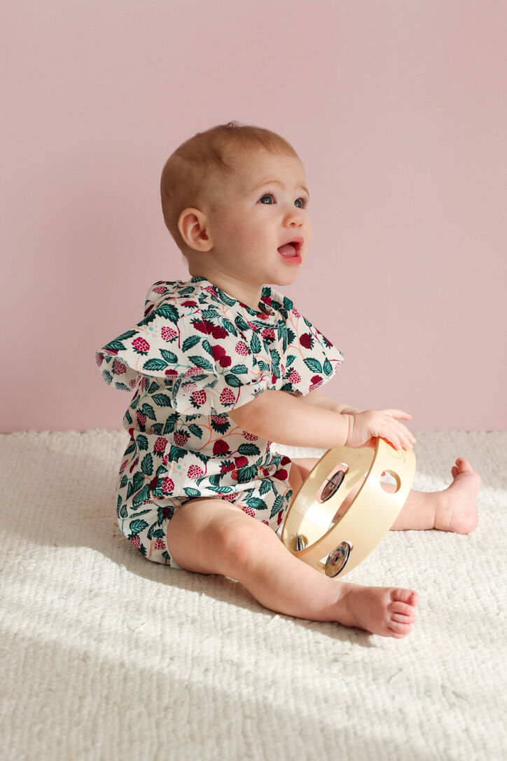 Baby in a floral outfit holding a tambourine against a pink background