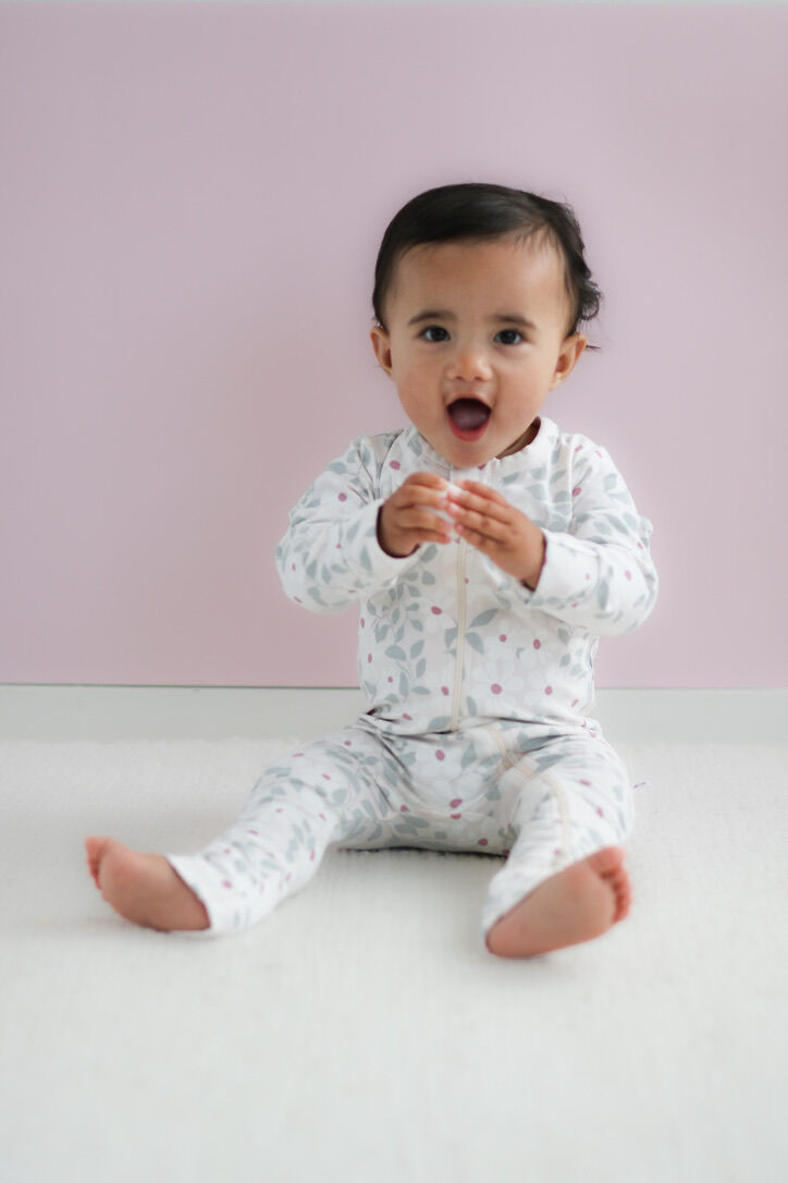 Baby sitting on a white surface wearing a white onesie with small patterns against a light pink background