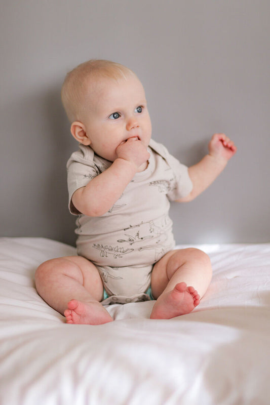 Baby sitting on a bed wearing a light-colored onesie.
