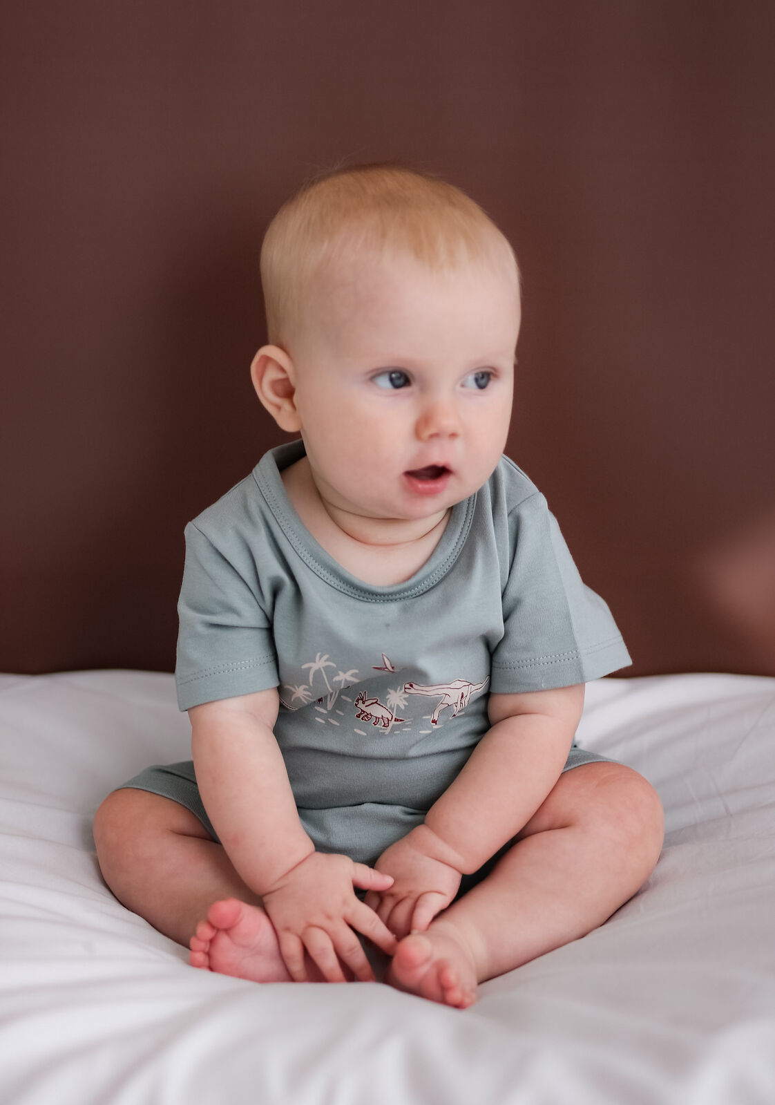 Baby sitting on a bed wearing a light gray onesie with a design.