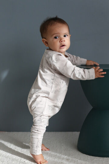 Baby in a light gray onesie standing next to a dark green stool against a gray wall.